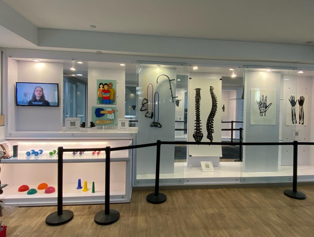 A view of of display cases. There is a monitor with a woman speaking. Below are a variety of colorful devices, cones, balls, weights, used in physical therapy. there are two colorful pop-ups of children, a boy in a cast, and a leg. next to it are a pair of small shoes connected by a rod. On the wall hang more physical therapy devices. There are black papercuts on white paper of two views of a spine, two views of the hand and forearm. and a hand.
