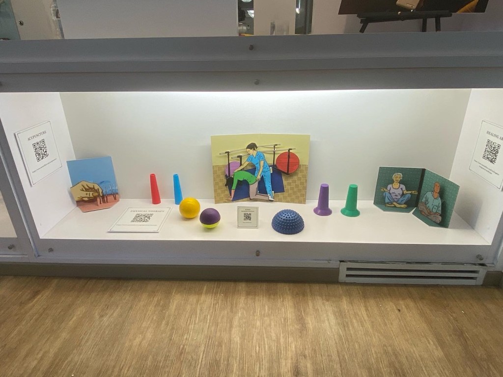 A display case with colorful popups of a hand administering accupuncture, a woman in blue scrubs in a physical therapy room assisting another woman who is stretching her spine, and two older individuals meditating. The paper pieces are surrounded by colorful physical therapy devices, cones, balls.