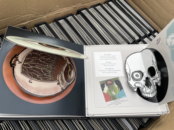 A pop-up book spread sits open on a case of books. they feature the interior of the eye and a black and white pop-up of a skull with an eye in its socket.
