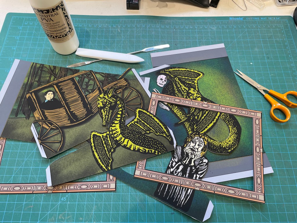 A green cutting mat with bone folder, glue, and scissors. A stack of cut papers.