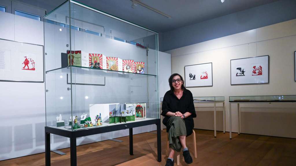 Woman with brown hair and glasses sits in a chair in a room surrounded by paper cuts and pop-up books.